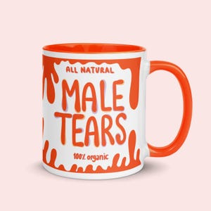 May include: A white ceramic mug with an orange interior and handle. The exterior features orange liquid drip patterns and the text "ALL NATURAL MALE TEARS 100% organic" printed in orange with a light purple shadow effect on the lettering.