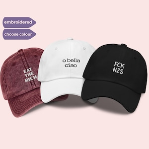 May include: Three baseball caps in maroon, white, and black. The maroon cap features white embroidered text reading EAT THE RICH. The white cap has black embroidered text reading o bella ciao. The black cap has white embroidered text reading FCK NZS.