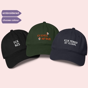 May include: Three embroidered baseball caps in black, dark green, and navy blue. The black cap has "FCK NZS" embroidered in white. The green cap has "ICH SCHEISS AUF NAZIS" with bird graphics. The navy cap reads "KEIN MENSCH IST ILLEGAL" in white.