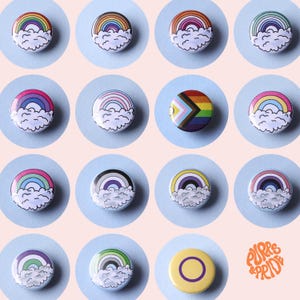 Peut inclure: Une collection de boutons ronds colorés avec des motifs d'arc-en-ciel et des nuages. Les boutons présentent divers drapeaux de la fierté, dont l'arc-en-ciel traditionnel, les drapeaux transgenres et non binaires. Les boutons sont disposés sur un fond rose clair.
