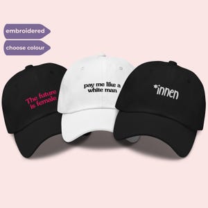 Può includere: Tre cappellini da baseball con testo ricamato. Il cappellino a sinistra è nero con testo rosa che recita 'The future is female'. Il cappellino centrale è bianco con testo nero che recita 'pay me like a white man'. Il cappellino a destra è nero con testo bianco che recita '*innen'.