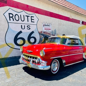 Könnte beinhalten: Ein roter Chevrolet Bel Air Cabriolet aus dem Jahr 1955, das vor einem Gebäude mit einem auf die Wand gemalten Route 66-Schild geparkt ist. Das Auto hat ein weißes Verdeck und goldene Verzierungen.