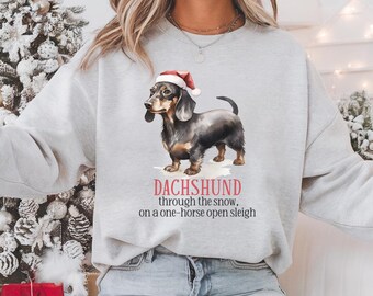 Peut inclure: Un sweat-shirt gris avec un teckel de Noël portant un chapeau de Père Noël. Le texte sur le sweat-shirt est "Dachshund through the snow, on a one-horse open sleigh."