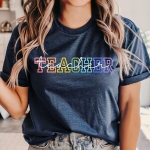 Puede incluir: Una camiseta azul marino con la palabra "TEACHER" impresa en colores arcoíris. Las letras están delineadas en negro.