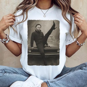 Könnte beinhalten: Weißes T-Shirt mit einem Sepia-Bild einer Person in einem schwarzen Overall und Maske, die sich auf ein Auto stützt, ein Bein angehoben. Das Shirt ist ein lässiger Artikel für den täglichen Gebrauch.
