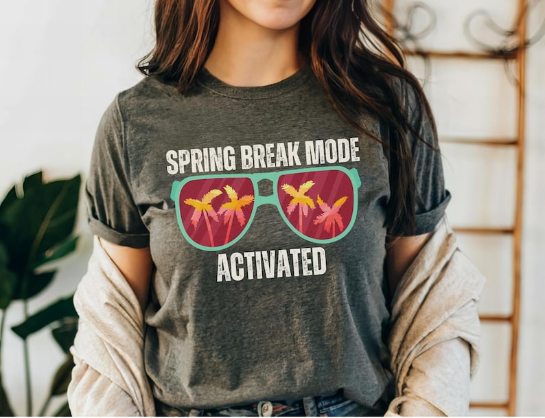 以下が含まれることがあります： 「SPRING BREAK MODE ACTIVATED」の文字が入った濃いグレーのTシャツ。グラフィックは、レンズの中にヤシの木が入ったサングラスが特徴です。サングラスは、ティールフレームと赤いレンズです。シャツはカジュアルなスタイルです。