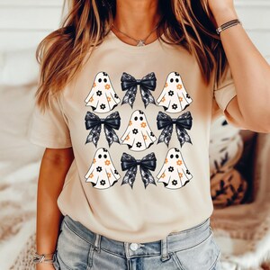 Puede incluir: Una camiseta beige con un patr&oacute;n de fantasmas y lazos negros. Los fantasmas son blancos con flores naranjas y los lazos son negros con encaje blanco.