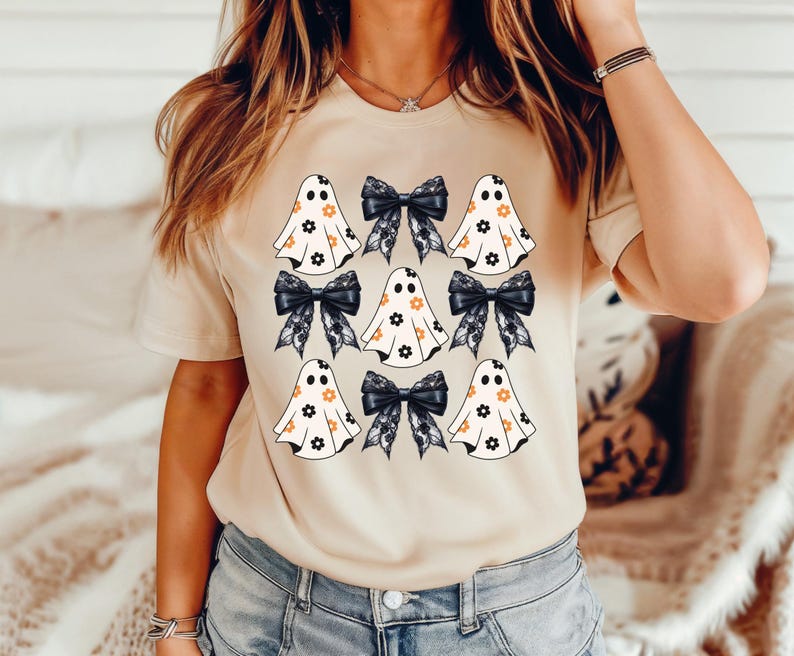 Puede incluir: Una camiseta beige con un patr&oacute;n de fantasmas y lazos negros. Los fantasmas son blancos con flores naranjas y los lazos son negros con encaje blanco.