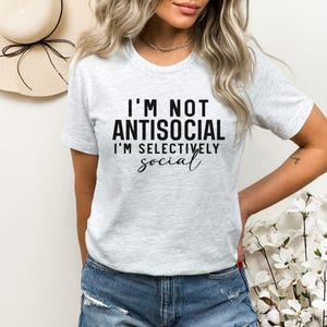Op de afbeelding: Een grijs T-shirt met de tekst "I'm not antisocial I'm selectively social" in zwart gedrukt.
