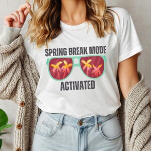 以下が含まれることがあります： 「SPRING BREAK MODE ACTIVATED」の文字が入った白いTシャツ。ヤシの木が描かれたサングラスのグラフィックが特徴です。カジュアルウェアアイテムです。