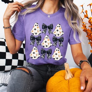 Puede incluir: Una camiseta morada con un patr&oacute;n de fantasmas blancos con lazos negros y dise&ntilde;os florales. Los fantasmas llevan lazos negros con ribete de encaje.