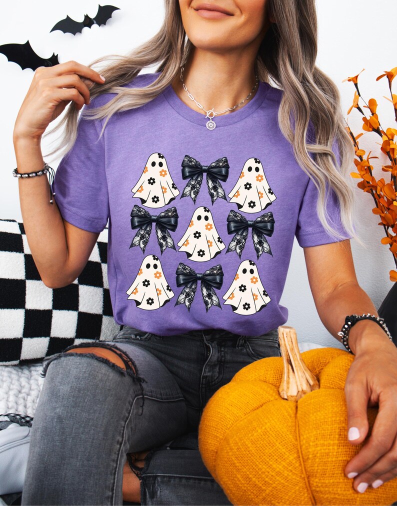 Puede incluir: Una camiseta morada con un patr&oacute;n de fantasmas blancos con lazos negros y dise&ntilde;os florales. Los fantasmas llevan lazos negros con ribete de encaje.