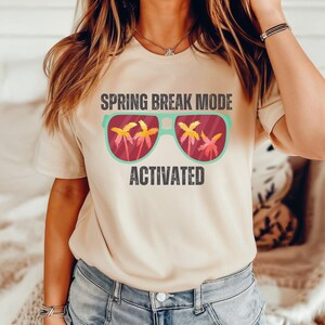 以下が含まれることがあります： 「SPRING BREAK MODE ACTIVATED」の文字が入ったベージュ色のTシャツ。グラフィックは、レンズの中にヤシの木が描かれたサングラスです。サングラスはティールフレームで、ヤシの木は黄色とオレンジ色です。