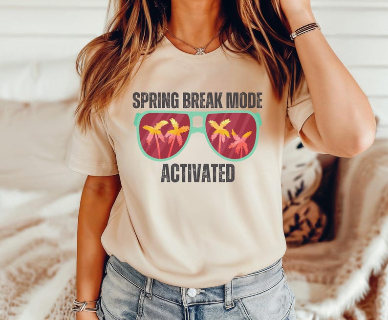 以下が含まれることがあります： 「SPRING BREAK MODE ACTIVATED」の文字が入ったベージュ色のTシャツ。グラフィックは、レンズの中にヤシの木が描かれたサングラスです。サングラスはティールフレームで、ヤシの木は黄色とオレンジ色です。