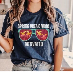 以下が含まれることがあります： 「SPRING BREAK MODE ACTIVATED」の文字が入ったネイビーブルーのTシャツ。グラフィックは、レンズにヤシの木が映ったサングラスが特徴です。このシャツは柔らかい素材で作られています。
