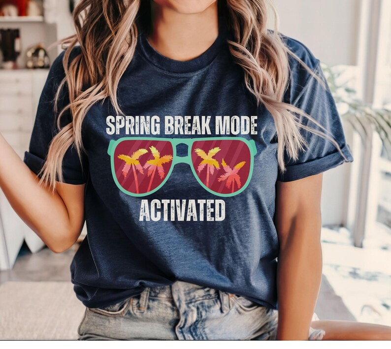 以下が含まれることがあります： 「SPRING BREAK MODE ACTIVATED」の文字が入ったネイビーブルーのTシャツ。グラフィックは、レンズにヤシの木が映ったサングラスが特徴です。このシャツは柔らかい素材で作られています。