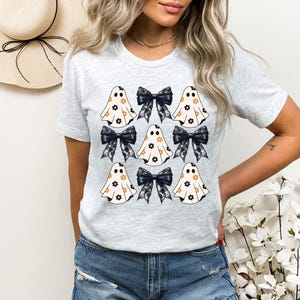 Puede incluir: Una camiseta gris con un patr&oacute;n de fantasmas blancos con flores naranjas y lazos negros. Los fantasmas est&aacute;n dispuestos en una cuadr&iacute;cula.