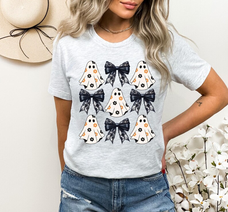Puede incluir: Una camiseta gris con un patr&oacute;n de fantasmas blancos con flores naranjas y lazos negros. Los fantasmas est&aacute;n dispuestos en una cuadr&iacute;cula.
