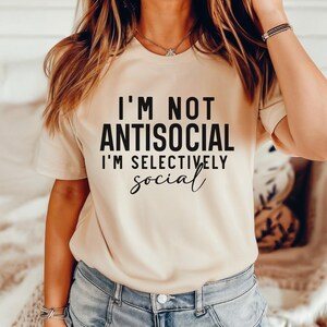 Op de afbeelding: Een beige T-shirt met de tekst "I'm not antisocial I'm selectively social" in zwart gedrukt.