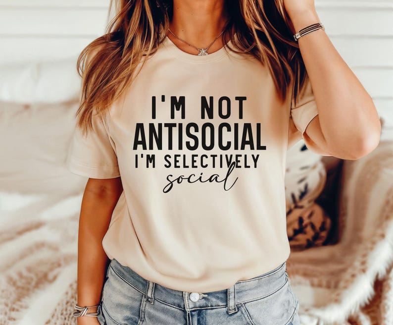 Op de afbeelding: Een beige T-shirt met de tekst "I'm not antisocial I'm selectively social" in zwart gedrukt.
