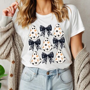 Puede incluir: Camiseta blanca con un patr&oacute;n de fantasmas blancos y negros con flores naranjas y lazos de encaje negro.