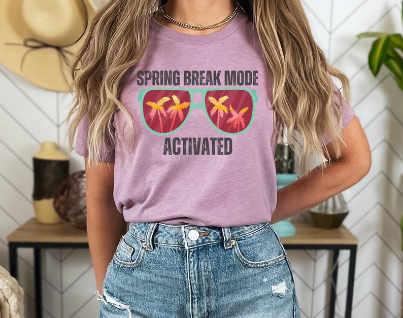 以下が含まれることがあります： ヤシの木が描かれたサングラスの上に「SPRING BREAK MODE ACTIVATED」の文字がプリントされたライラック色のTシャツ。青いデニムのショートパンツと合わせています。