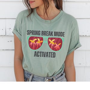 以下が含まれることがあります： セージグリーンのTシャツ。テキストは「SPRING BREAK MODE ACTIVATED」。グラフィックは、レンズにヤシの木が映ったサングラスです。カジュアルで快適なスタイルです。