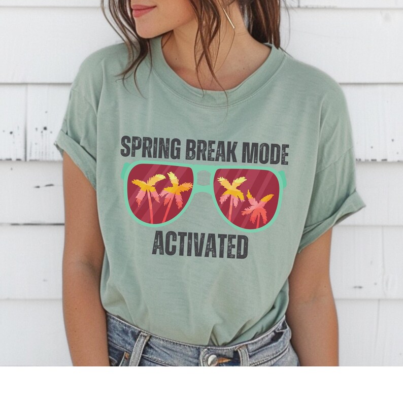 以下が含まれることがあります： セージグリーンのTシャツ。テキストは「SPRING BREAK MODE ACTIVATED」。グラフィックは、レンズにヤシの木が映ったサングラスです。カジュアルで快適なスタイルです。