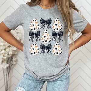 Puede incluir: Una camiseta gris con un estampado gr&aacute;fico de varios fantasmas blancos con lazos negros y patrones florales. Los fantasmas tienen ojos negros y una peque&ntilde;a sonrisa.