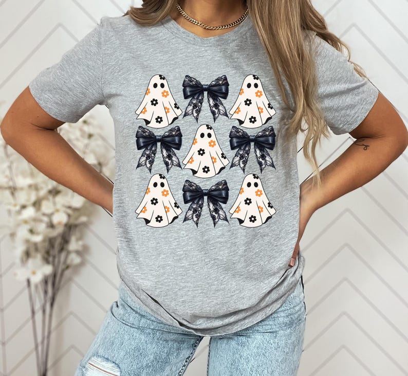 Puede incluir: Una camiseta gris con un estampado gr&aacute;fico de varios fantasmas blancos con lazos negros y patrones florales. Los fantasmas tienen ojos negros y una peque&ntilde;a sonrisa.