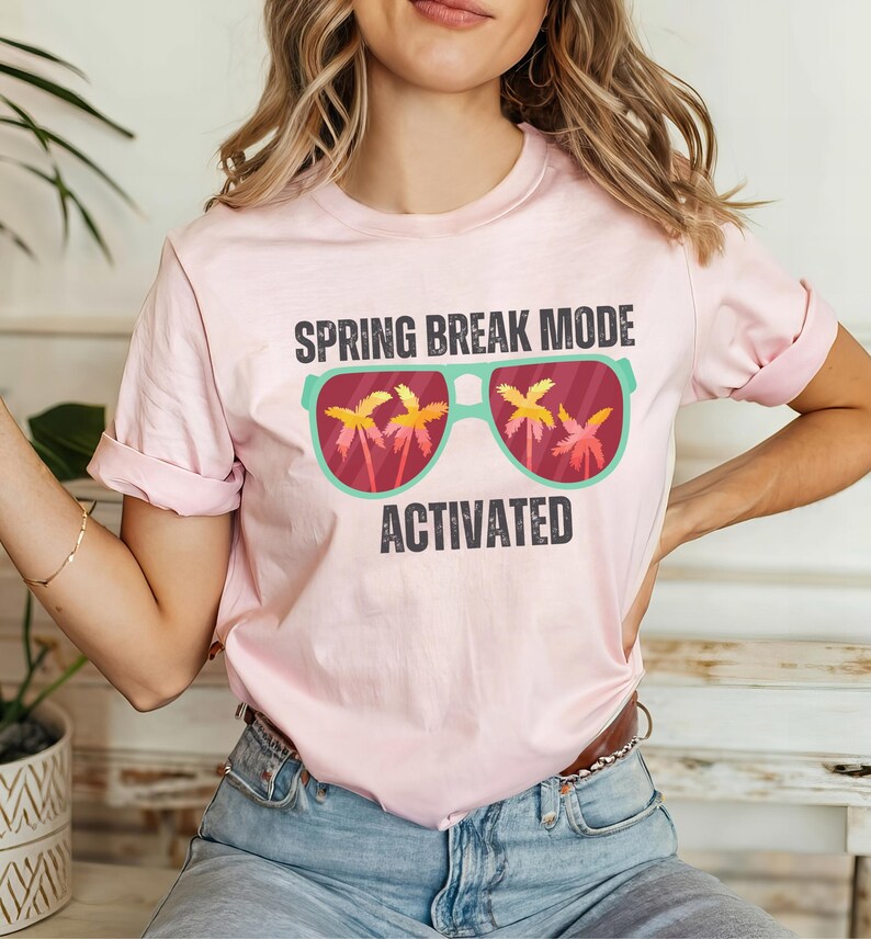 以下が含まれることがあります： 袖をまくった薄いピンク色のTシャツ。ヤシの木のグラフィックと「SPRING BREAK MODE ACTIVATED」の文字が黒とティールでプリントされています。
