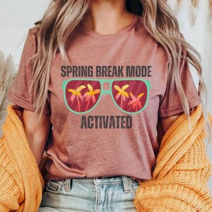 以下が含まれることがあります： 「SPRING BREAK MODE ACTIVATED」の文字が入ったモーヴ色のTシャツ。ヤシの木が入ったサングラスのグラフィック。青いジーンズと黄色のセーターを着ている人。