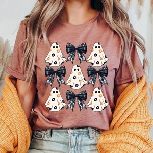 Puede incluir: Una camiseta malva con un estampado gr&aacute;fico de fantasmas con patrones florales y lazos negros.