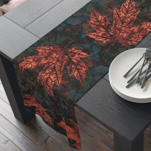 May include: A dark brown wooden table with a fall-themed table runner featuring orange and red maple leaves on a blue and green background. The table runner is set with two wine glasses, a plate with cutlery, and a bowl of apples.