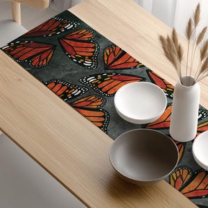 May include: A wooden dining table with a butterfly-themed table runner, two white bowls, and a gray bowl. A white vase with dried plants sits on the table. Two wooden chairs with gray seats are positioned on either side of the table.