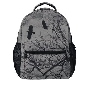 May include: A grey backpack featuring a black silhouette print of two birds flying over bare tree branches. The bag includes a top handle, a front zippered pocket, and black mesh side pockets on both sides for storage of various items.