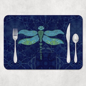 May include: A rectangular placemat featuring a dark blue background with a central dragonfly illustration in shades of blue and green. A silver fork is positioned on the left, while a silver knife and a silver spoon are placed on the right side.
