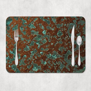 May include: A rectangular placemat featuring a mottled, oxidized copper-like pattern in shades of deep brown and verdigris green. A silver-toned metal fork is positioned on the left, while a silver-toned knife and spoon sit on the right side.