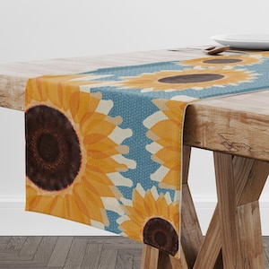 May include: A table runner with a sunflower design. The runner has a blue background with yellow sunflowers and a brown centre. The runner is placed on a wooden table.