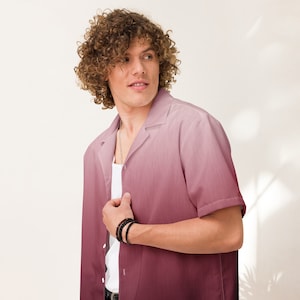 May include: A man with curly brown hair wearing a pink and white ombre button-up shirt with a white undershirt. He is wearing a silver chain necklace and black bracelets on his wrist.