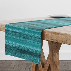 May include: Teal and black abstract patterned table runner. The runner is displayed on a wooden table with a white plate. The runner's design features horizontal brushstrokes of teal and black, creating a textured appearance. This table runner is a decorative home accent.