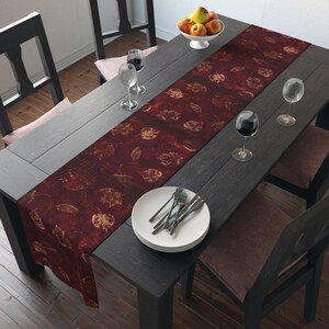 May include: A dark wooden dining table set for a meal. A burgundy table runner with a leaf pattern runs down the centre. White plates with forks and wine glasses are set. A bowl of apples sits at the table's end.