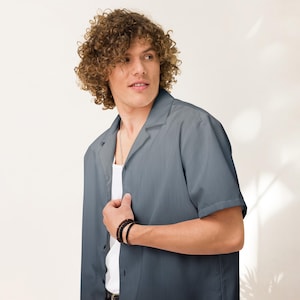 Puede incluir: Una camisa azul pizarra de manga corta con cuello de campamento, usada sobre una camiseta interior blanca. La camisa está abotonada en la parte superior y tiene un ajuste relajado. Una pulsera negra es visible en la muñeca.