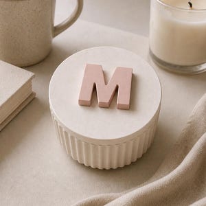 May include: A beige ceramic container with a pink letter "M" on top, next to a mug of coffee, a candle in a glass, and a small book. The container has a ribbed base and a flat, round lid. The background is a neutral color.