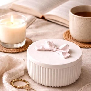 May include: A white, round trinket box with a ridged base and a decorative bow on the lid. A lit candle in a glass, a cup of tea, a gold beaded bracelet, and an open book are also visible.