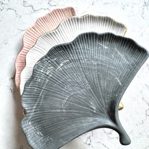 Op de afbeelding: Drie decoratieve, blad-vormige keramische borden in een waaierarrangement. De borden zijn roze, wit en grijs, met gouden randen. De borden hebben een gestructureerd oppervlak en liggen op een marmeren achtergrond.