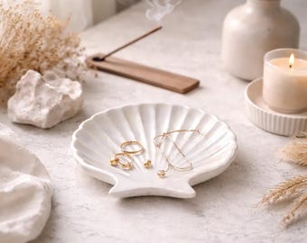 Puede incluir: Un plato de joyería blanco en forma de concha contiene anillos, pendientes y un delicado collar dorados. Una vela encendida, un jarrón con flores secas e incienso completan la serena escena de tonos neutros. El humo se eleva del incienso.