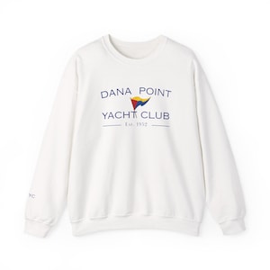 Könnte beinhalten: Ein weißes Sweatshirt mit Rundhalsausschnitt, das den blauen Schriftzug DANA POINT YACHT CLUB, eine kleine rot-gelb-blaue Wimpelgrafik und den Text Est. 1952 in der Mitte zeigt. Die Buchstaben YC sind in kleiner blauer Schrift auf dem rechten Ärmel.