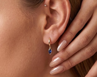 May include: A gold hoop earring featuring a small, marquise-cut blue gemstone charm dangling from the bottom. The earring is worn in the earlobe, and a small silver-toned stud is visible in the upper cartilage area of the ear.