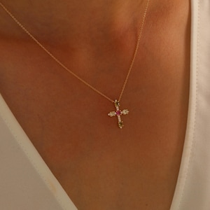 Könnte beinhalten: Eine goldene Kettenkette mit einem kleinen Kreuzanhänger, der mit bunten Edelsteinen verziert ist. Das Kreuz hat einen rosafarbenen Mittel-Stein, der von kleineren weißen, grünen und gelben Steinen umgeben ist.